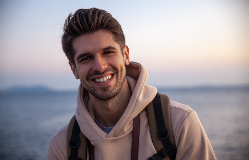A young man wearing a beige hoodie and backpack smiles outdoors near a body of water at sunset.