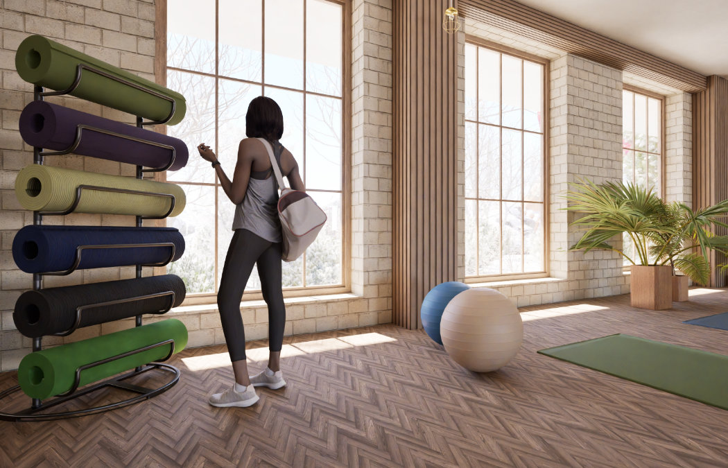 A woman in workout clothes stands by a rack of rolled yoga mats in a bright exercise studio with large windows and wood accents.